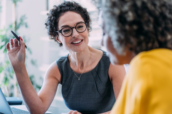 A white woman with short dark hair, weatring a grey top and glasses is holding a pen and sitting at a laptop is talking to a woman who is side on to the camera and wearing a yello top
